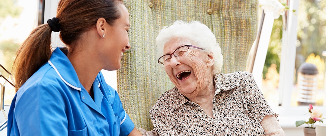 Nurse in blue scrubs and an elderly woman sharing a hearty laugh together outside.